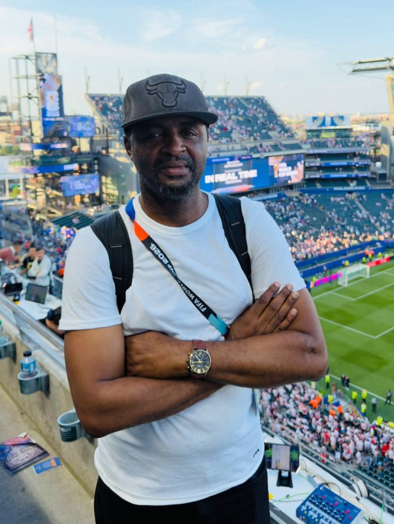 A man with a black Bulls cap and a white t-shirt standing with arms crossed in a stadium, with a large crowd and a soccer field visible in the background.