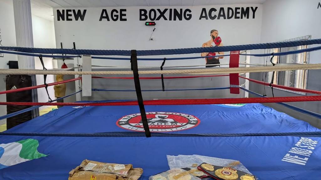 View of the interior of New Age Boxing Academy, featuring a boxing ring with red and blue ropes, a blue mat, and championship belts on the floor. A mural of a boxer is visible on the wall.
