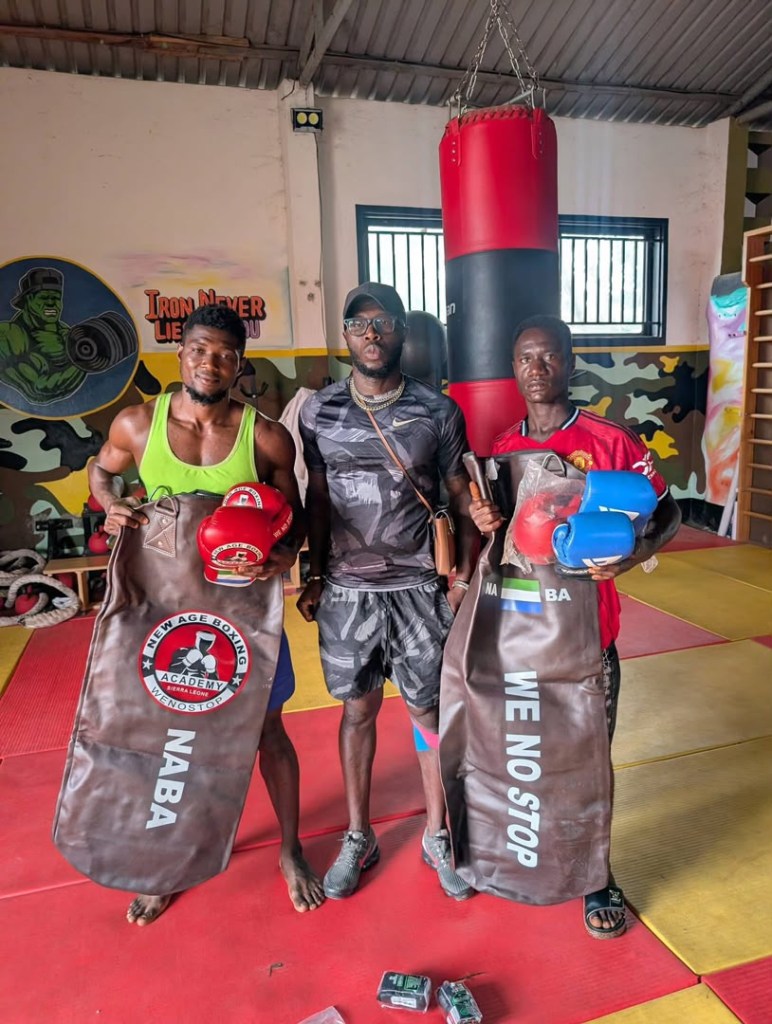 Three men in a boxing gym, holding training gear and wearing boxing gloves, posing in front of a punching bag. One man wears a green tank top, another a black jersey, and the third a red shirt. The gym has a vibrant mural and various training equipment.