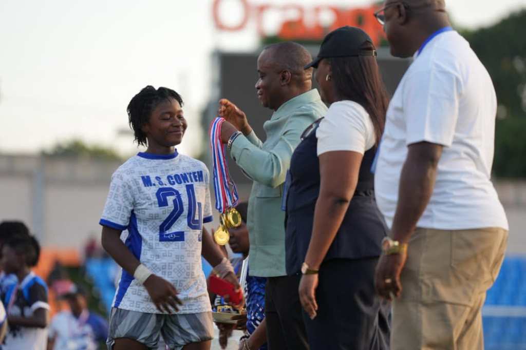 A young female athlete in a sports jersey receiving a medal and awards from an official during a ceremony, with other attendees in the background.