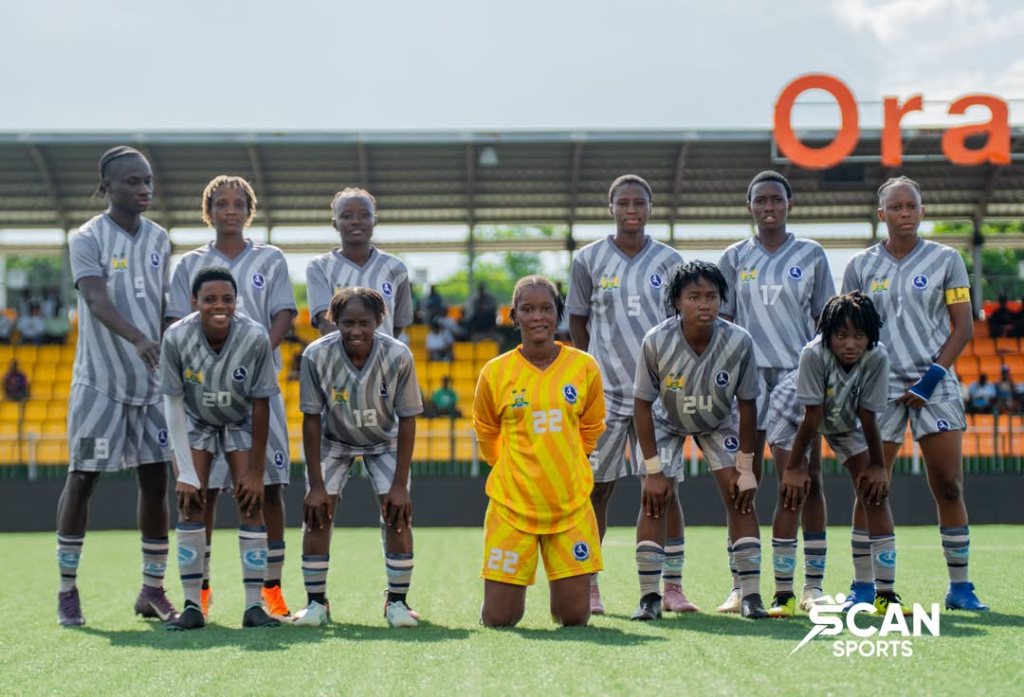 A women's soccer team poses for a group photo on the field, wearing gray and yellow uniforms. The team consists of eleven players, with one goalkeeper in bright yellow, and the others in matching gray jerseys. The setting features an outdoor stadium with spectators in the background.